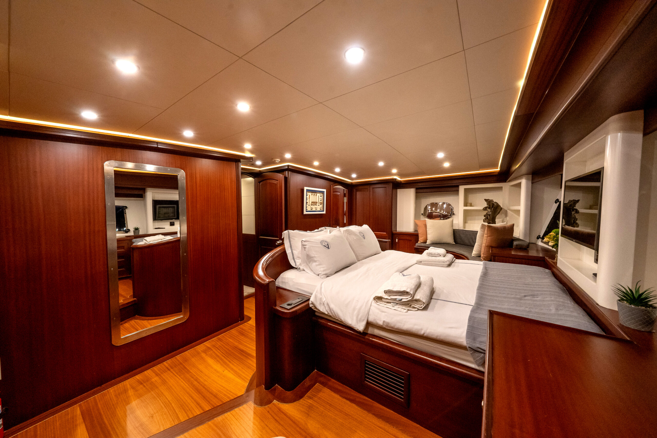 VIRTUOSO Luxurious yacht bedroom with polished wood walls and floors, a large bed with white linens, built-in shelves, soft lighting, a full-length mirror, and decorative items, creating a spacious and elegant atmosphere.