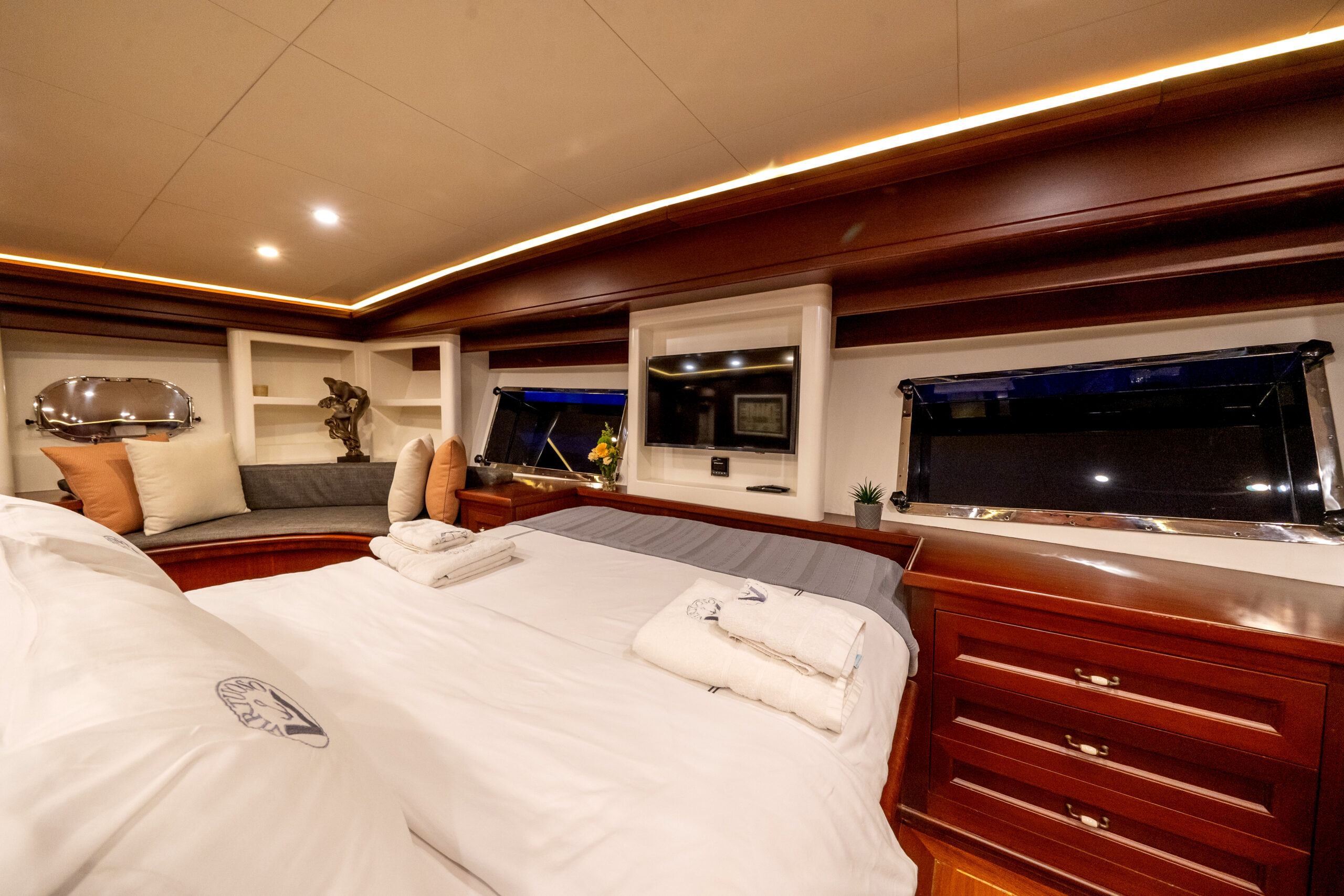 VIRTUOSO Luxurious yacht bedroom with a large bed, plush white linens, wood-paneled walls, windows, drawers, a cushioned seating area with pillows, towels on the bed, and soft recessed lighting.
