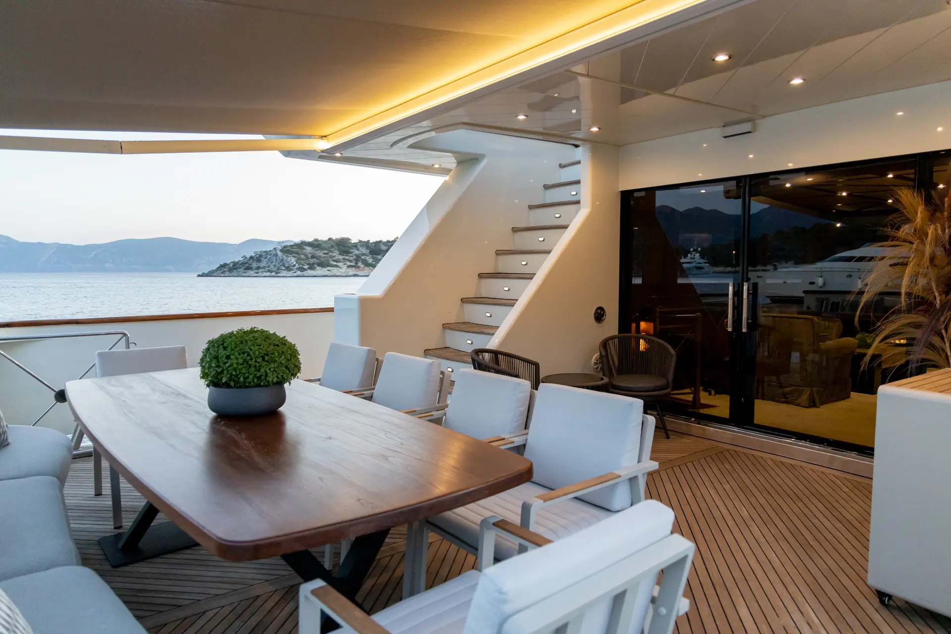 A luxury yacht deck with a wooden dining table, cushioned chairs, stairs leading up, and sliding glass doors to an indoor lounge. Ocean and mountains are visible in the background.
