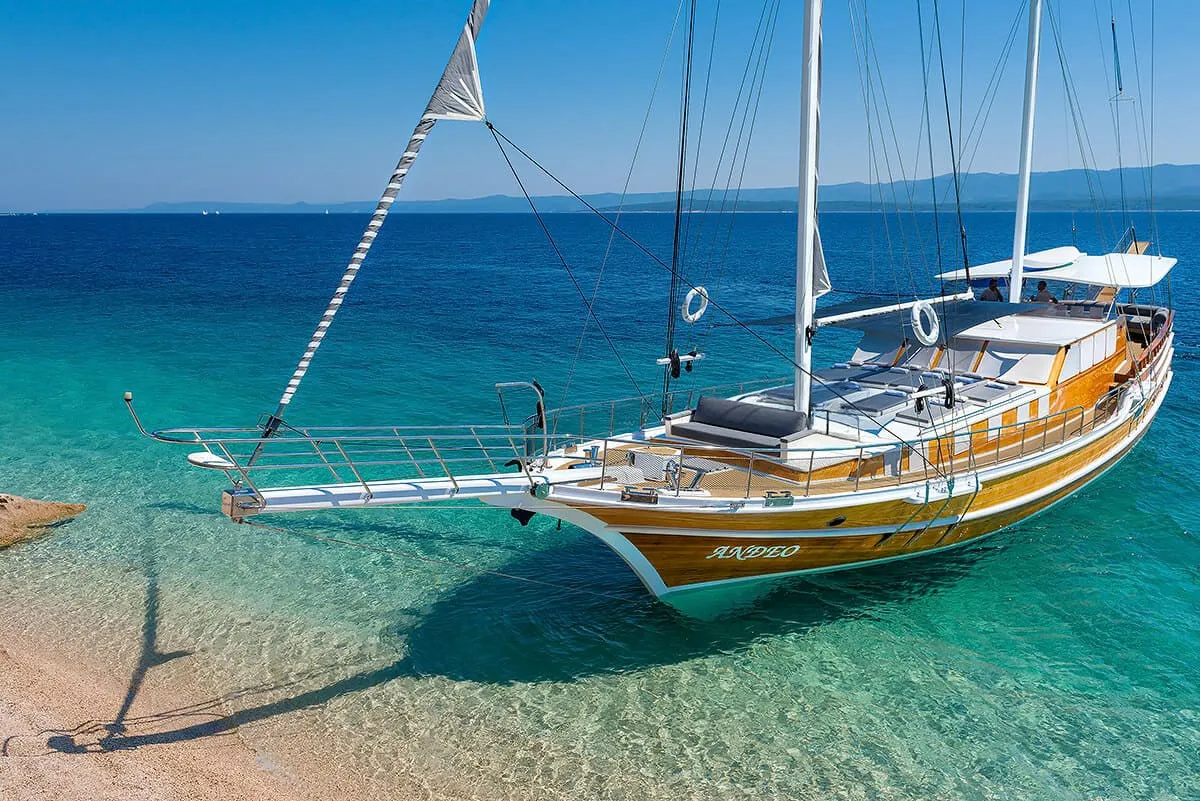 A wooden yacht with white sails is anchored close to a sandy beach in clear turquoise waters under a bright blue sky, with mountains visible in the distance.