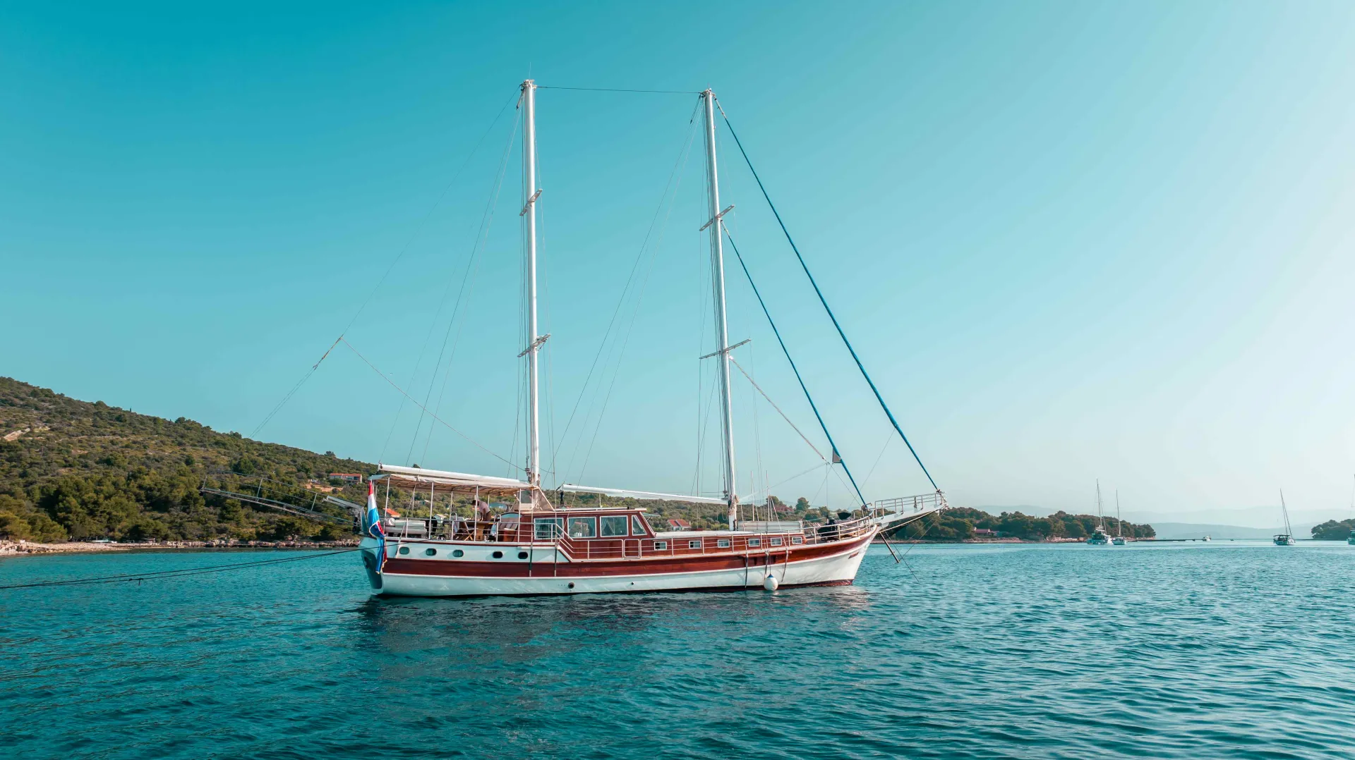 A large white and red sailing boat with two masts floats on calm blue water near a green, hilly shoreline under a clear blue sky.