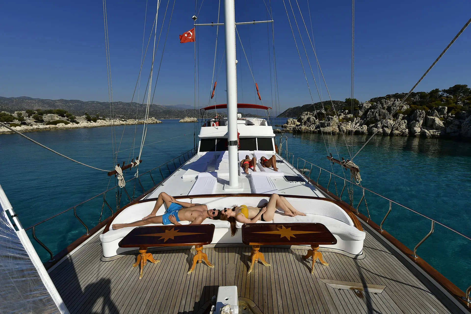 Two people sunbathe on the deck of a yacht in clear blue water, with rocky shores and greenery in the background under a clear sky. A Turkish flag flies at the stern.