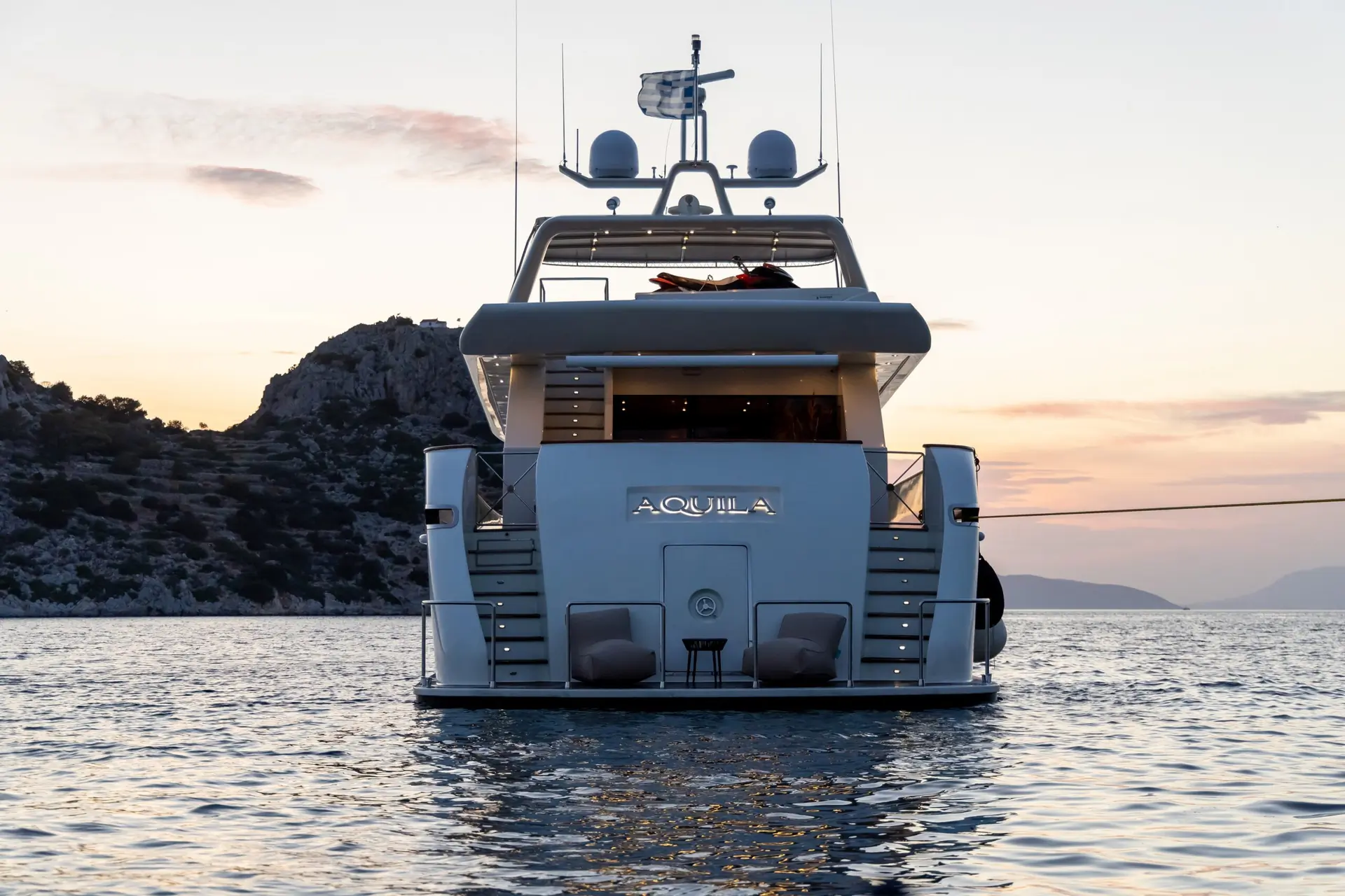 A white luxury yacht named “AQUILA” is anchored on calm water at sunset, with two chairs and a table on its deck. Mountains are visible in the background under a pastel sky.