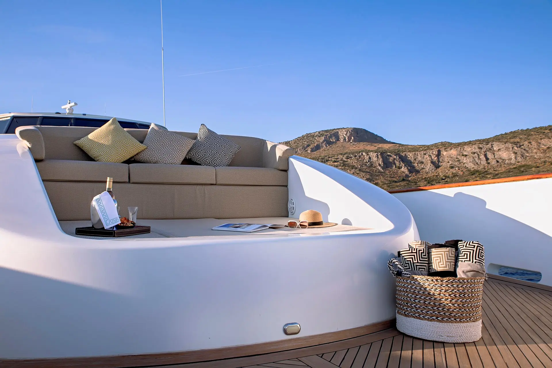 A cozy yacht seating area with beige cushions, a tray holding a drink and snacks, magazines, a hat, and a woven basket, set against a backdrop of rocky hills and clear blue sky.