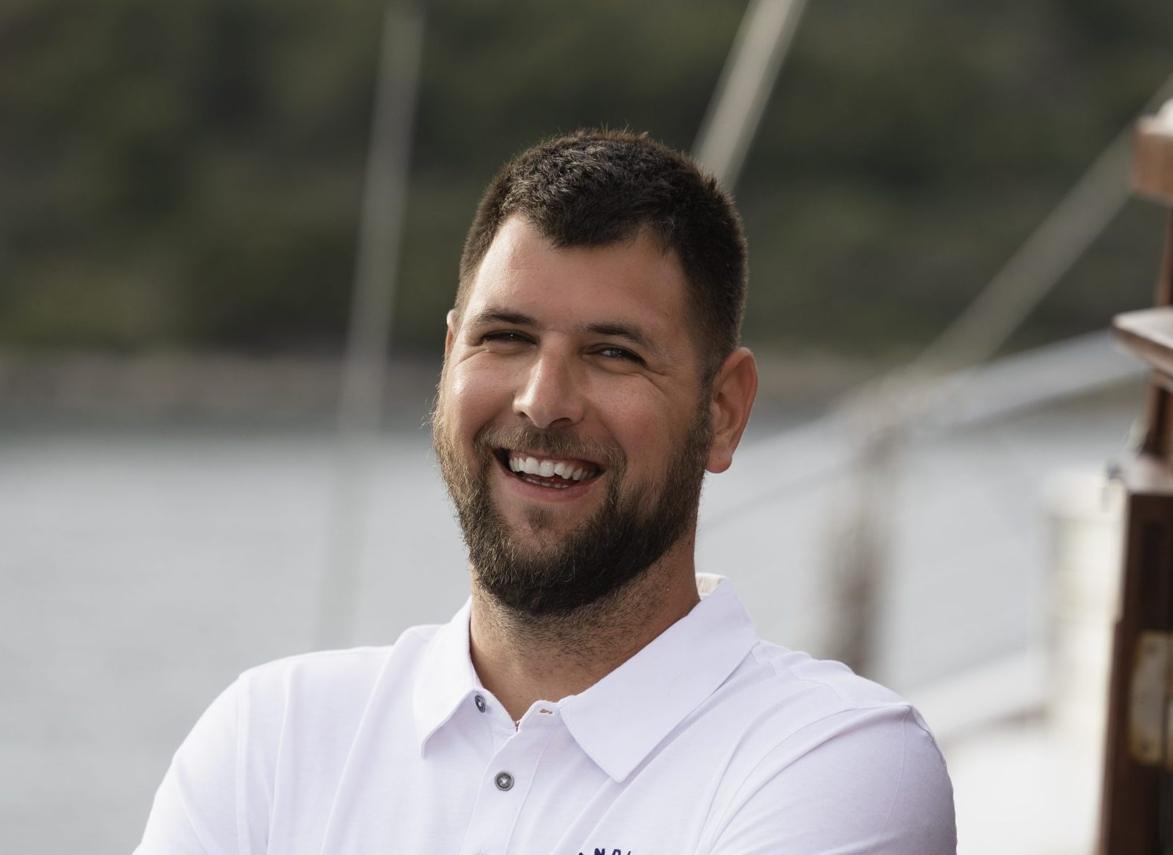 A man with short dark hair and a beard, wearing a white polo shirt with a sailboat logo, stands smiling with arms crossed on the deck of a boat near the water.