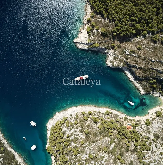 Aerial view of a blue-green bay surrounded by rocky, forested coastline, with several boats anchored in the water near the shore. The word Cataleya is overlaid on the image.