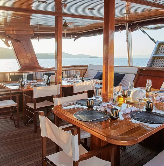 Elegant dining area on a wooden yacht deck set with tables, chairs, glassware, and placemats, overlooking calm water and distant hills under a clear sky.
