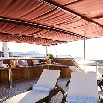 A luxurious yacht deck with cushioned lounge chairs, tables set for breakfast, rolled towels, and drinks, shaded by a red canopy. The ocean and mountains are visible in the background.