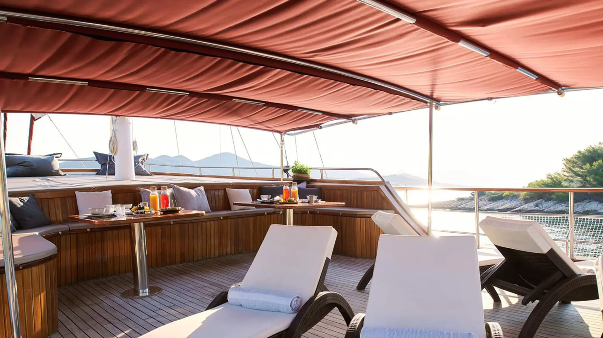 Luxury yacht deck with cushioned lounge chairs, a shaded canopy, wooden seating area, and a table set for breakfast. The sea and distant hills are visible in the background on a sunny day.