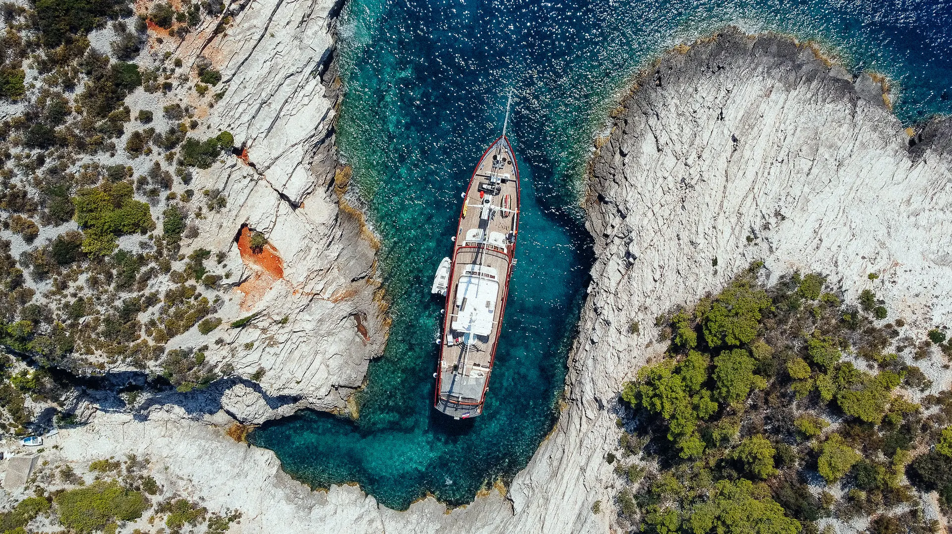 Aerial view of a boat anchored in a narrow, turquoise inlet surrounded by rocky cliffs with sparse greenery. The clear water reveals the boats shadow and underwater rocks.