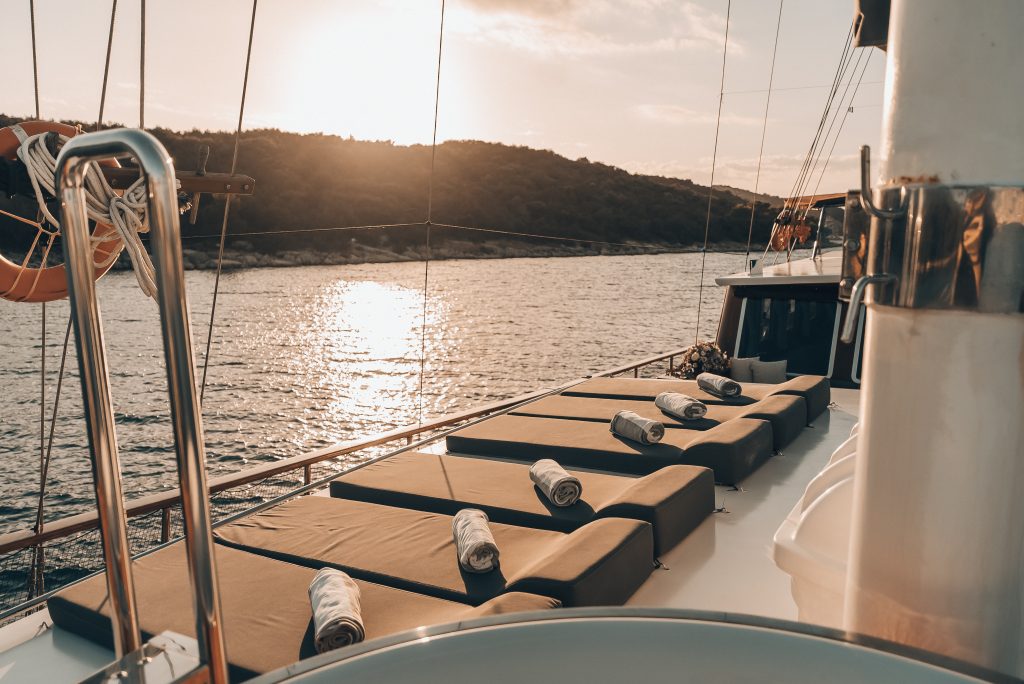 Sunset view from a yacht deck, featuring several cushioned lounge chairs with rolled towels, overlooking calm water and a tree-covered shoreline.