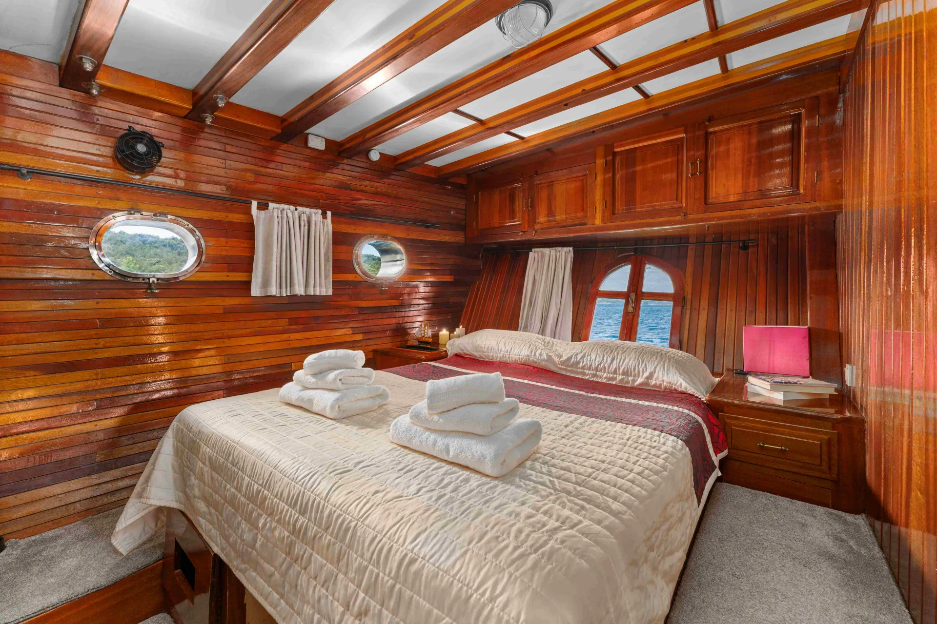 A cozy bedroom with polished wooden walls and ceiling, a double bed with folded towels on top, porthole windows, and a nightstand with a lamp, overlooking a view of water outside.