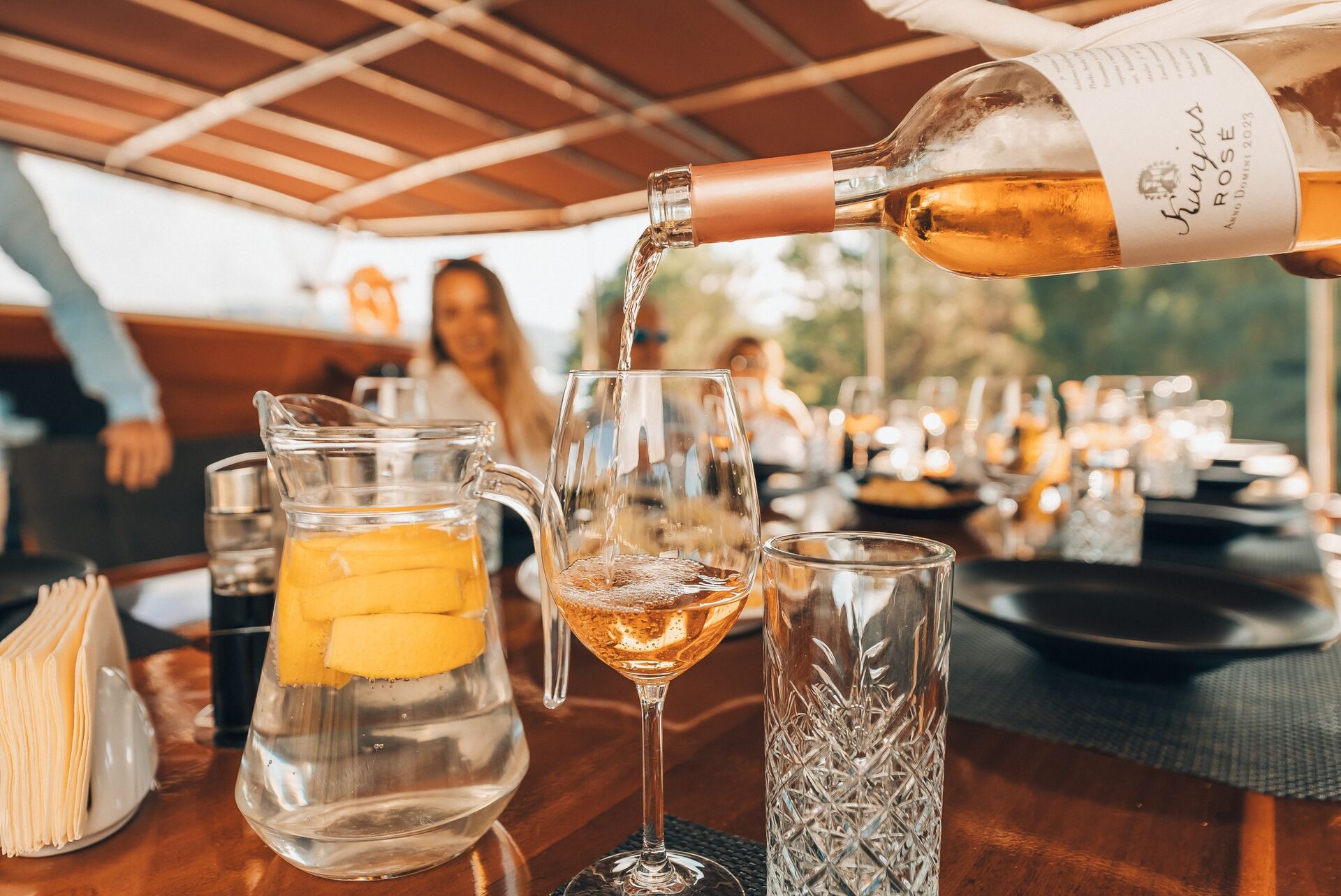 A close-up of rosé wine being poured into a glass on a set dining table with black plates, glassware, a pitcher of water with lemon slices, and blurred people in the background under a canopy.
