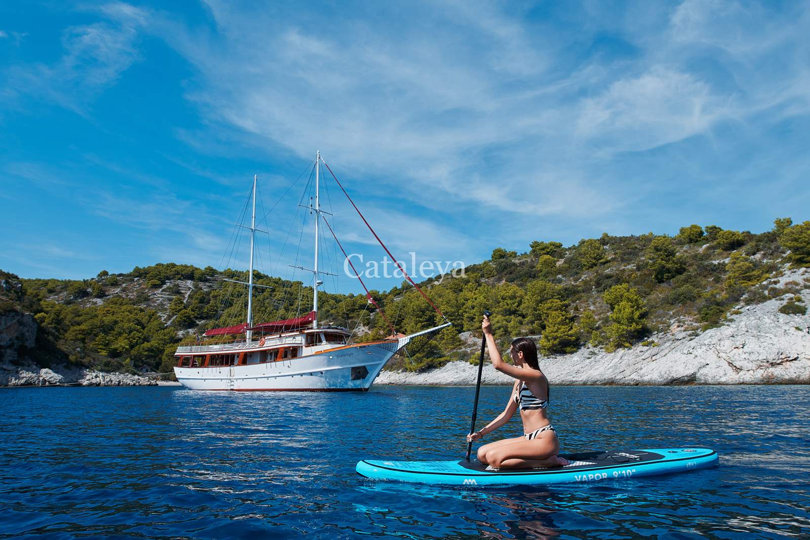 CATALEYA A woman in a swimsuit paddles on a blue stand-up paddleboard in calm blue water near a large white yacht, with a forested rocky shoreline and clear blue sky in the background.