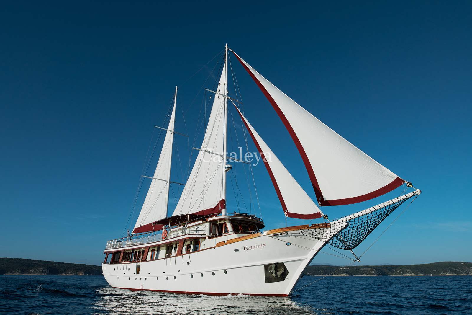 CATALEYA A large white sailing yacht with red trim sails on calm blue water under a clear sky. The yacht has two tall masts and multiple sails. Distant land and hills are visible on the horizon.