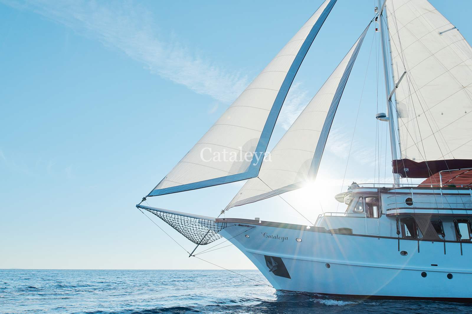 CATALEYA A white sailboat named Cataleya sails on calm blue water under a clear sky, with sunlight shining through its sails.