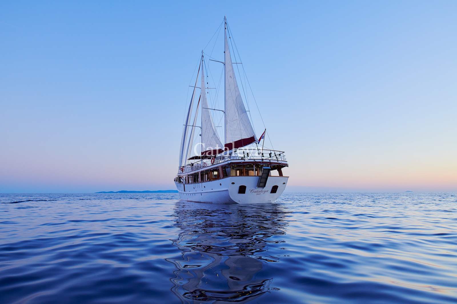 CATALEYA A white sailboat with raised sails floats on calm blue water during sunset, with a clear sky and distant land visible on the horizon.