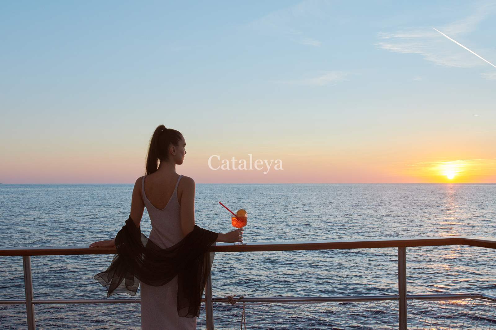 CATALEYA A woman in a white dress stands on a ship’s deck, holding a drink with a straw, looking out at the ocean during sunset. The sky is colorful, and the word Cataleya appears over the scene.