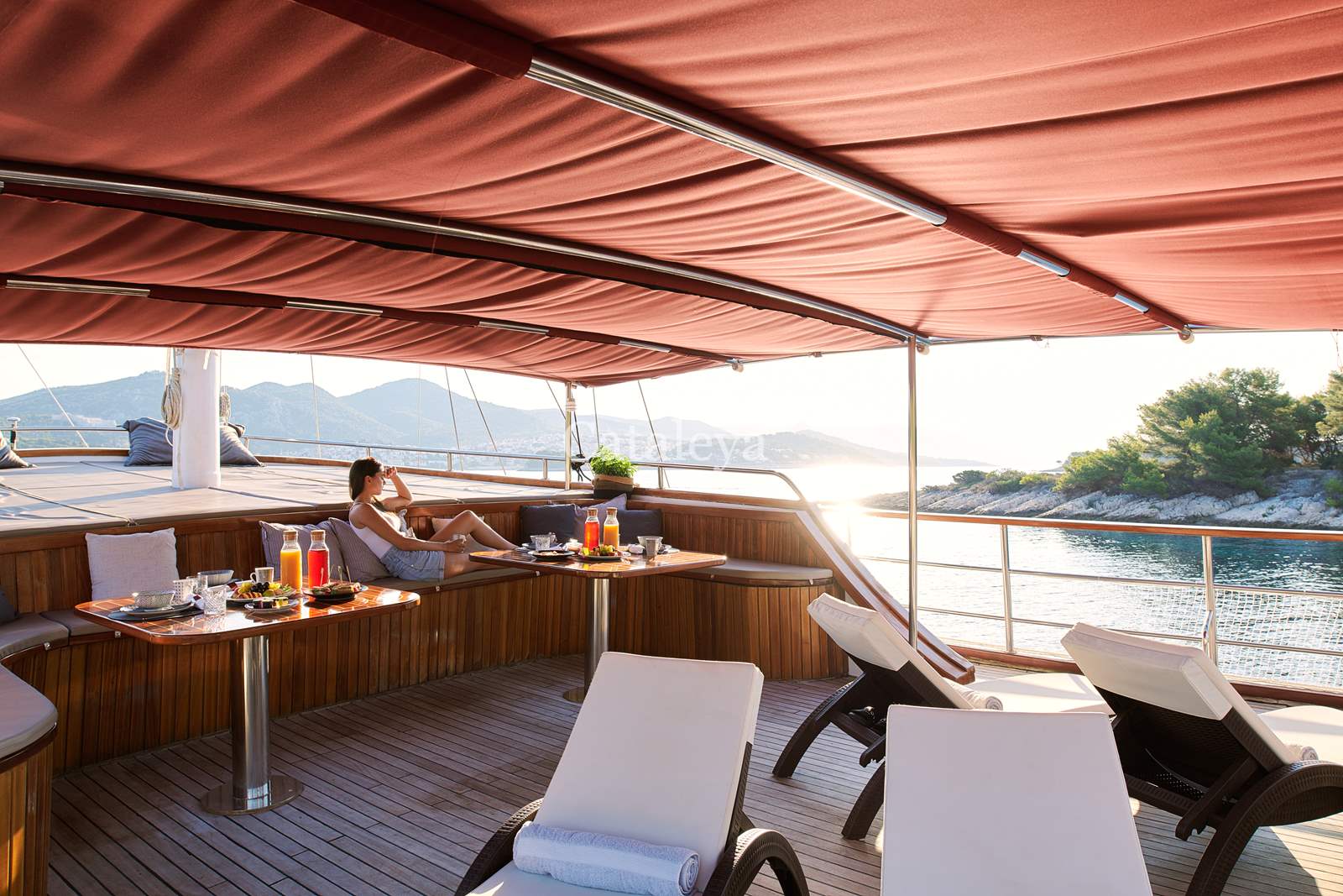 CATALEYA A woman relaxes on a lounge chair under a red canopy on a yacht, with breakfast items on a nearby table. The yacht is anchored near a scenic coastline with calm water and green hills in the background.