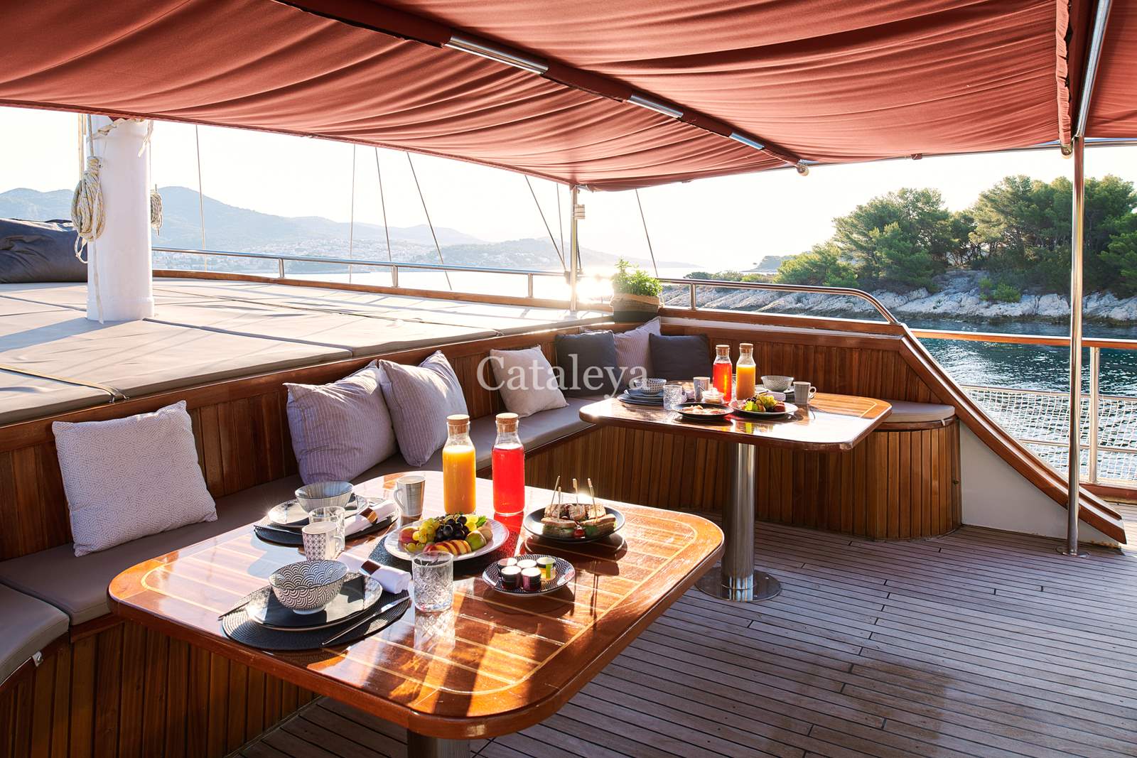 CATALEYA A shaded yacht deck features two wooden tables set with breakfast items, including juice, fruit, coffee, and pastries. Cushioned seating lines the railing, with scenic water and trees visible in the background.