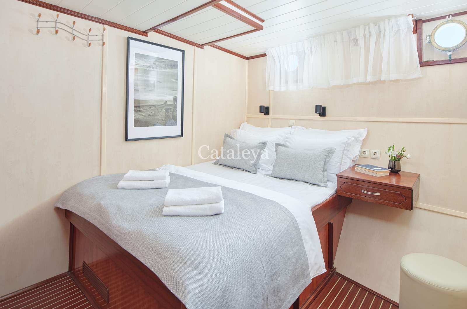 CATALEYA A cozy cabin with a double bed neatly made with white and gray linens, two sets of towels, a framed picture on the wall, a small wooden nightstand with flowers, and a round porthole window with white curtains.