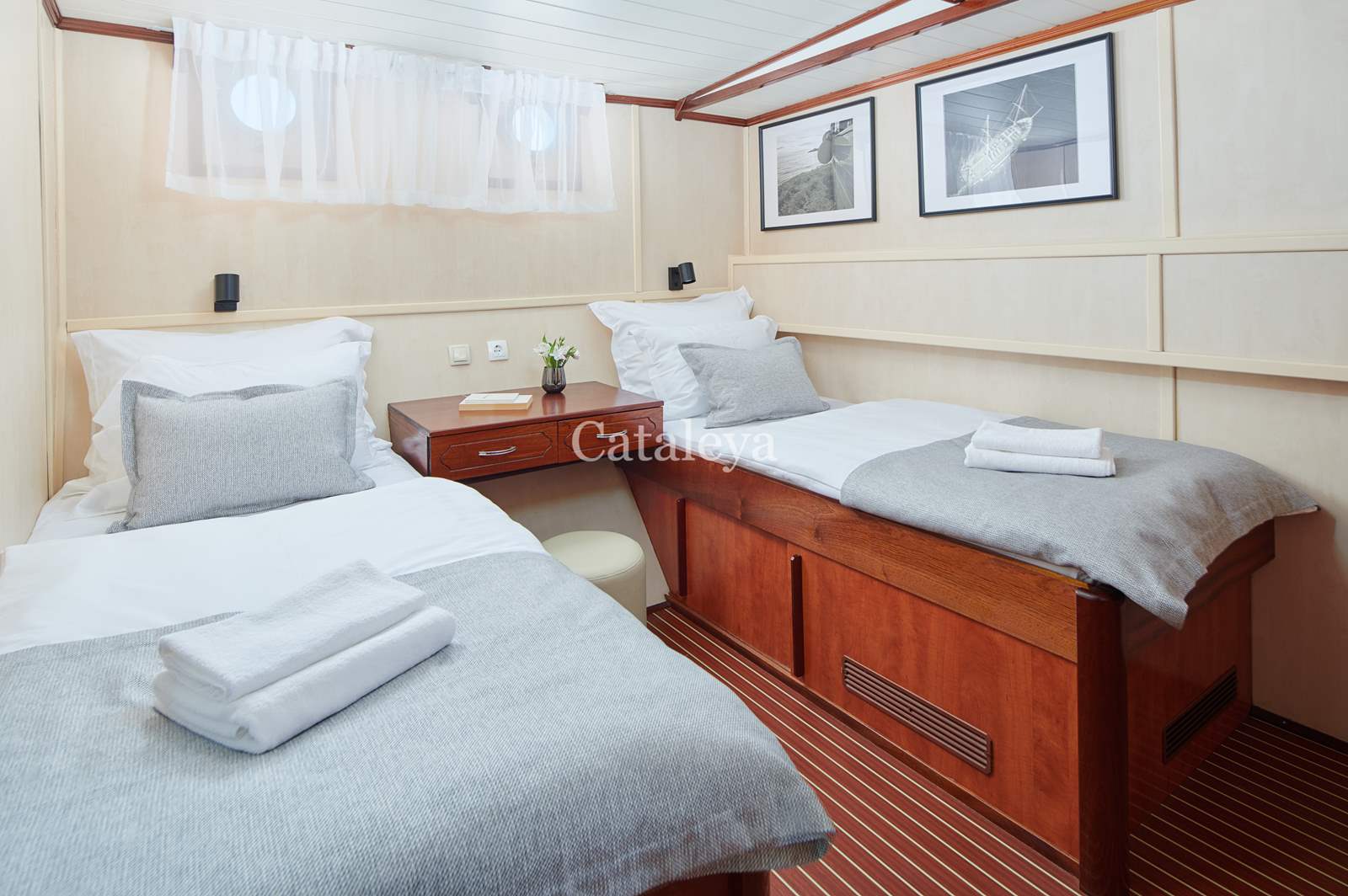 CATALEYA Cozy cabin room with two single beds, grey and white bedding, folded towels, a small central nightstand with a plant, and nautical-themed decor on the cream walls and striped wooden floor.