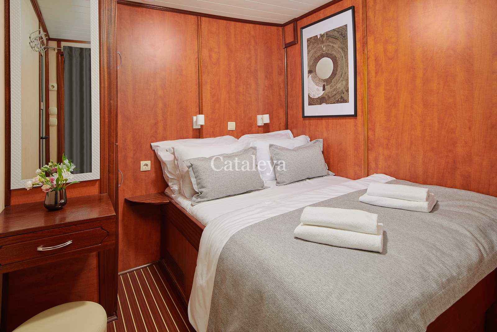 CATALEYA A cozy cabin bedroom with wooden walls, a double bed with grey and white bedding, two folded towels, a small vanity with a mirror and flowers, and a framed artwork above the bed.