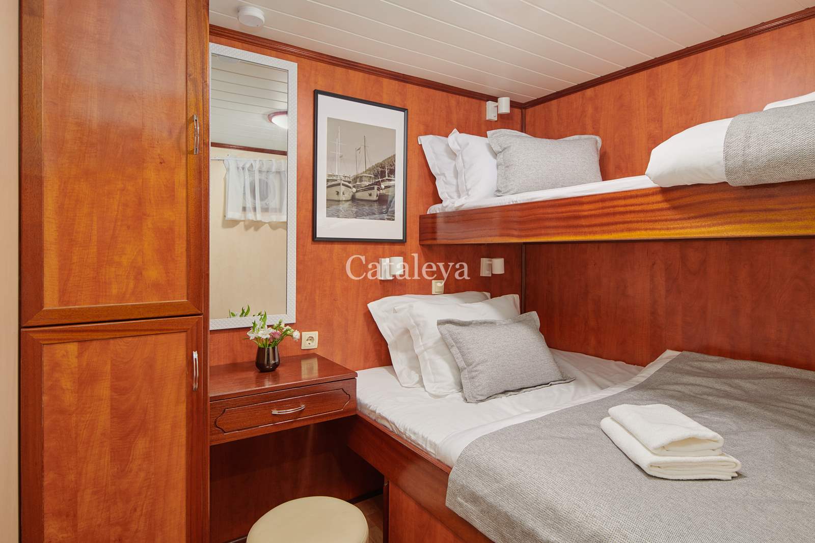 CATALEYA A cozy cabin room with wood-paneled walls features a bunk bed with white and gray bedding, a small desk with a flower vase, a stool, and a framed ship photo on the wall. Towels are folded neatly on the lower bed.