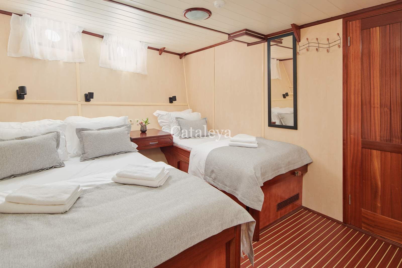 CATALEYA A cozy boat cabin with a double bed and a single bed, both with white and gray bedding. There is a wooden nightstand with flowers, a mirror on the wall, a wooden door, and striped flooring.