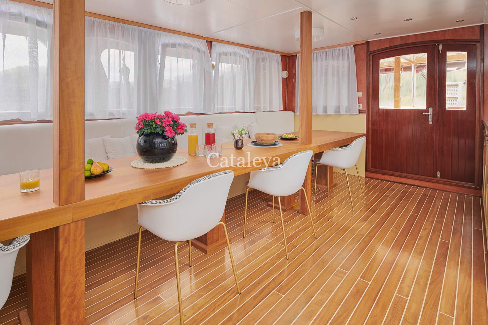 CATALEYA A bright, modern dining area with wood floors, a long wooden table, white chairs, potted flowers, fruit, and drinks. Sunlight streams through sheer curtains and a glass door at the back. Cataleya is written on the image.
