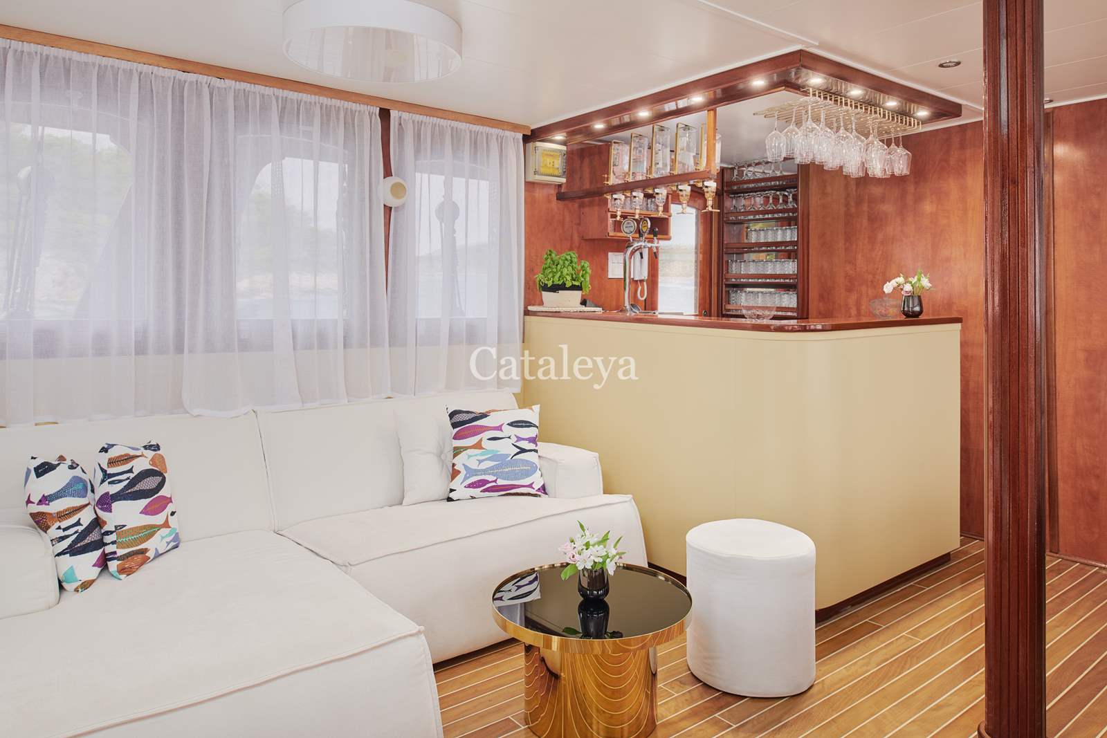 CATALEYA A cozy yacht lounge featuring a white sectional sofa with patterned pillows, a small round black coffee table with a flower vase, wood-paneled walls, a bar area with hanging glasses, and large windows with sheer curtains.