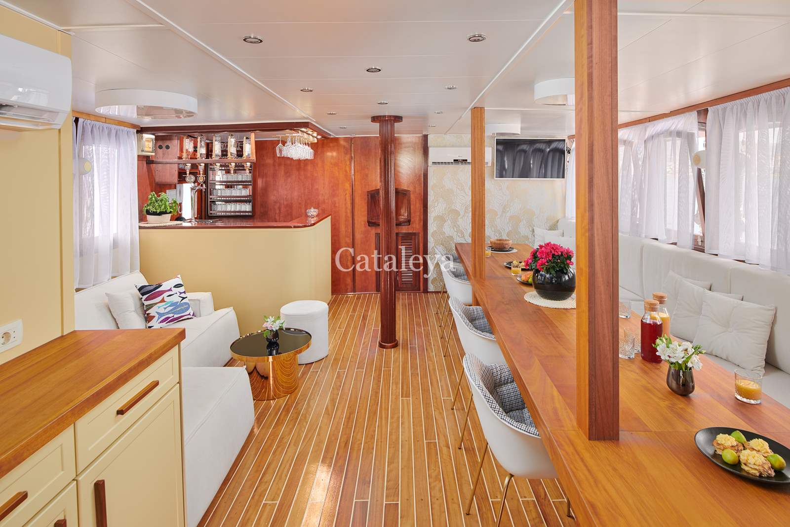 CATALEYA A bright, modern yacht interior with wood floors and walls, white sofas, a dining bar with stools, flowers, and large windows with white curtains. The space feels open and inviting, combining kitchen and lounge areas.