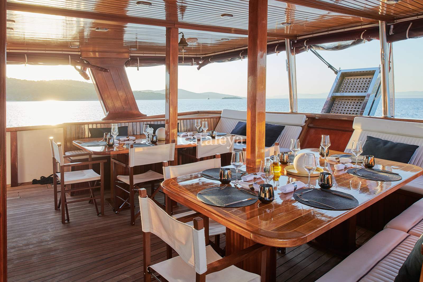 CATALEYA Elegant outdoor dining area on a wooden yacht, set with plates, glasses, and cutlery, overlooking calm sea and distant islands at sunset. The space has cushioned bench seating and natural light streaming in.