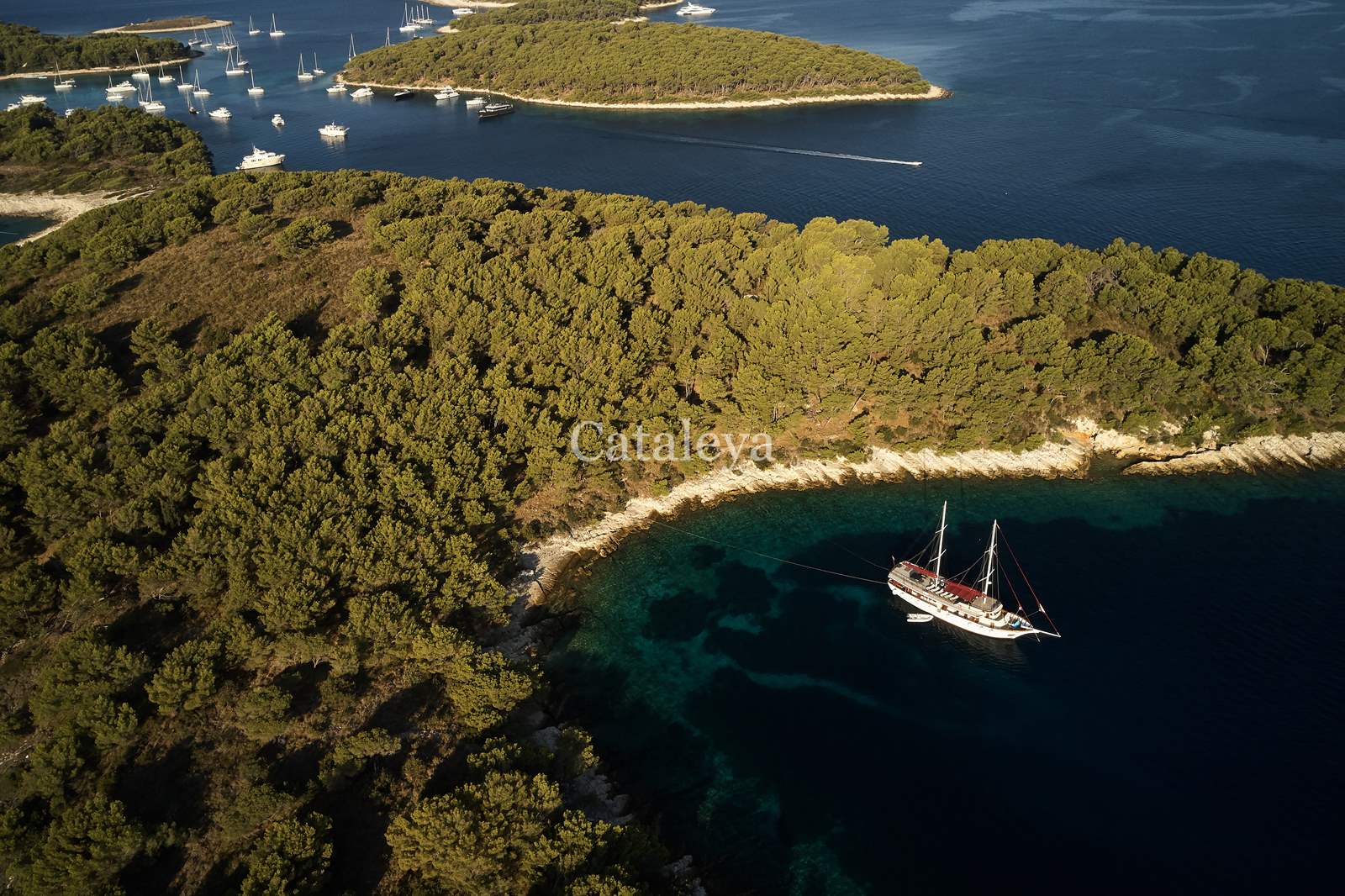 CATALEYA Aerial view of a sailboat anchored near a forested island with turquoise water, surrounded by several other islands and yachts in the distance under a clear sky.