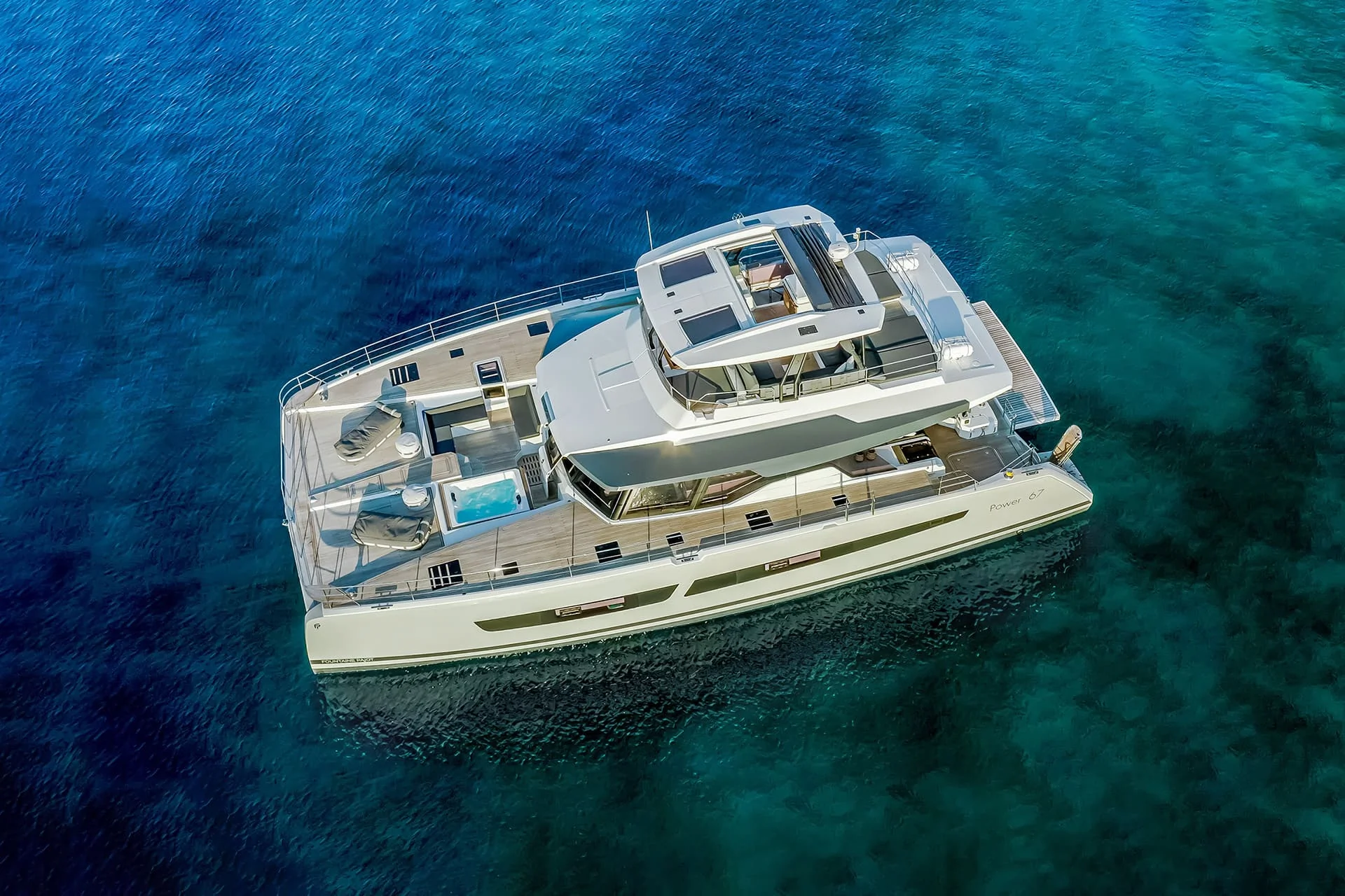 A luxury white catamaran yacht floats on clear blue water, viewed from above. The yacht has sunbeds, a seating area, and an upper deck, surrounded by calm, vibrant ocean.