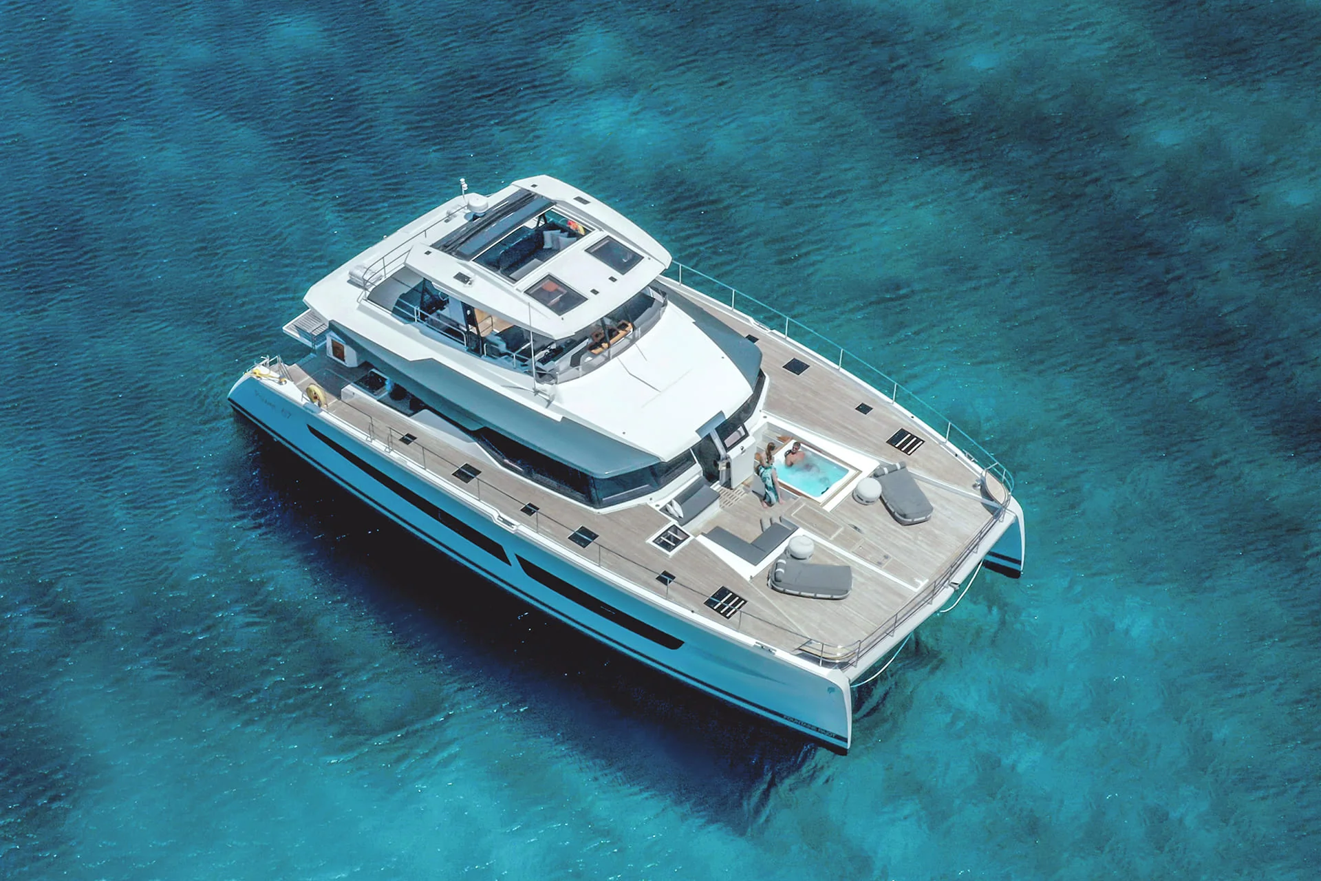 A modern white catamaran yacht floats on clear blue water, viewed from above. Two people relax in a small pool on the deck, surrounded by lounge chairs and open space.