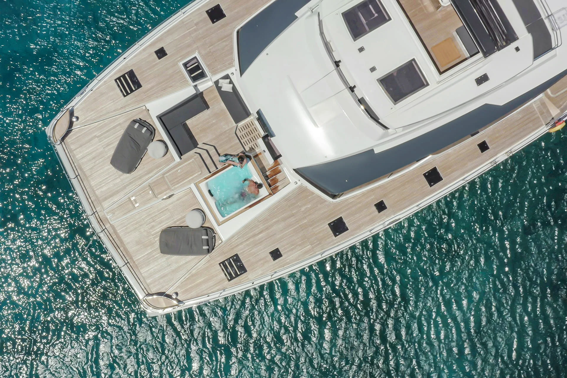 Aerial view of a luxury yacht on blue water, featuring a sun deck with lounge chairs and a couple relaxing in a small pool.
