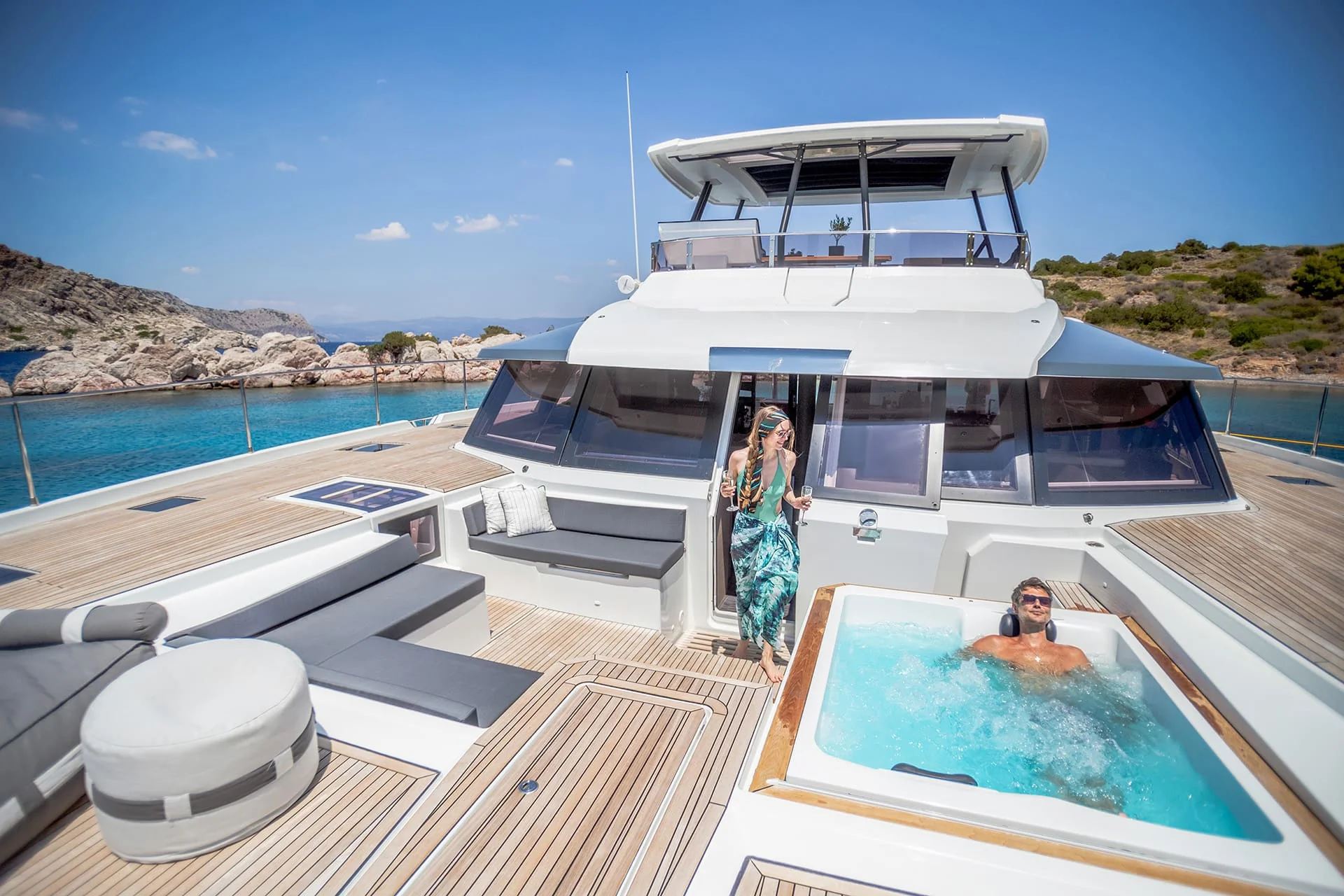 A woman stands at the doorway of a luxury yacht while a man relaxes in a hot tub on the deck. The yacht is anchored in clear blue water near a rocky coastline under a sunny sky.