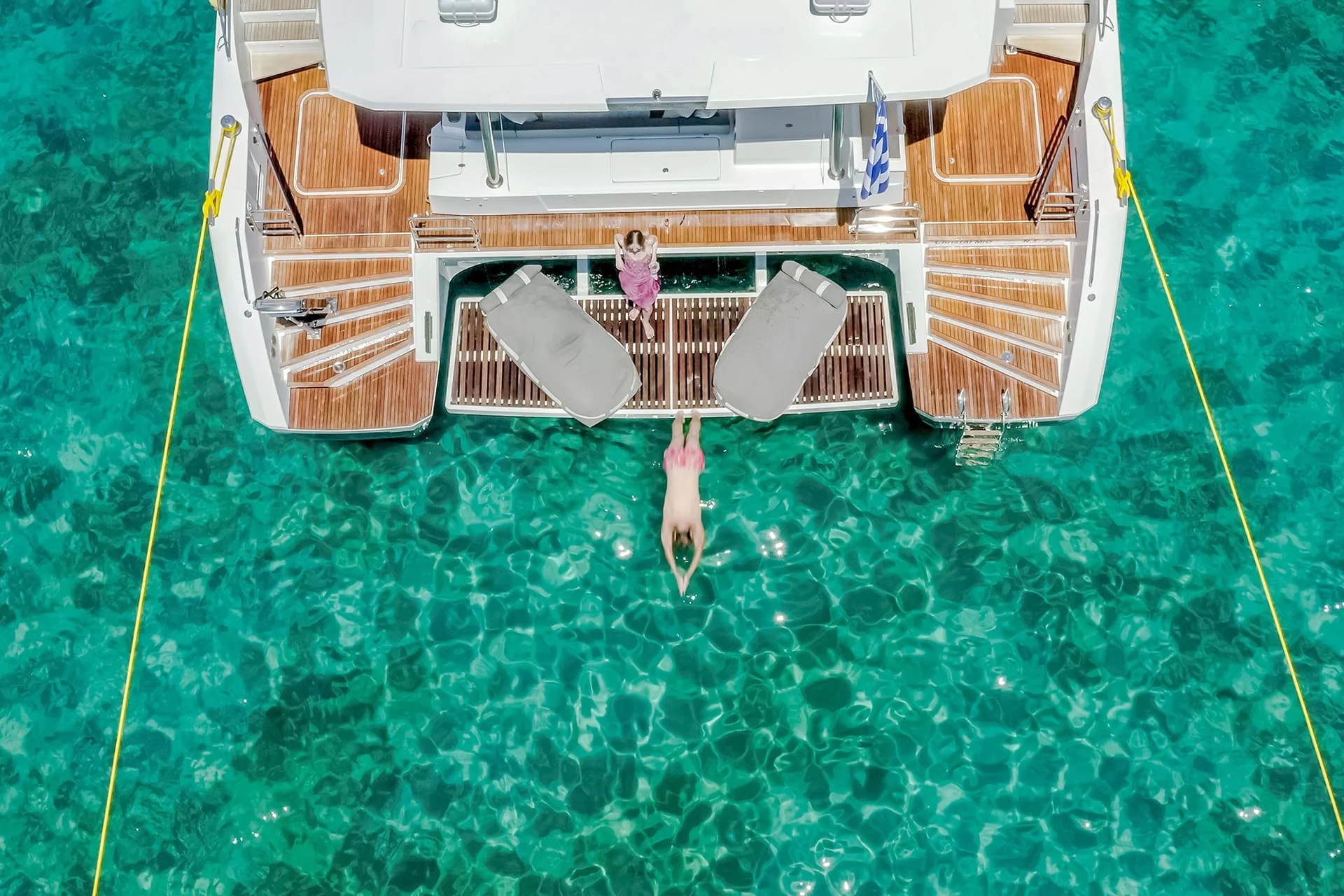 A person dives off the back of a yacht into clear turquoise water while another person stands nearby on the deck. The yacht has two lounge chairs at the rear, and the water below is bright and transparent.