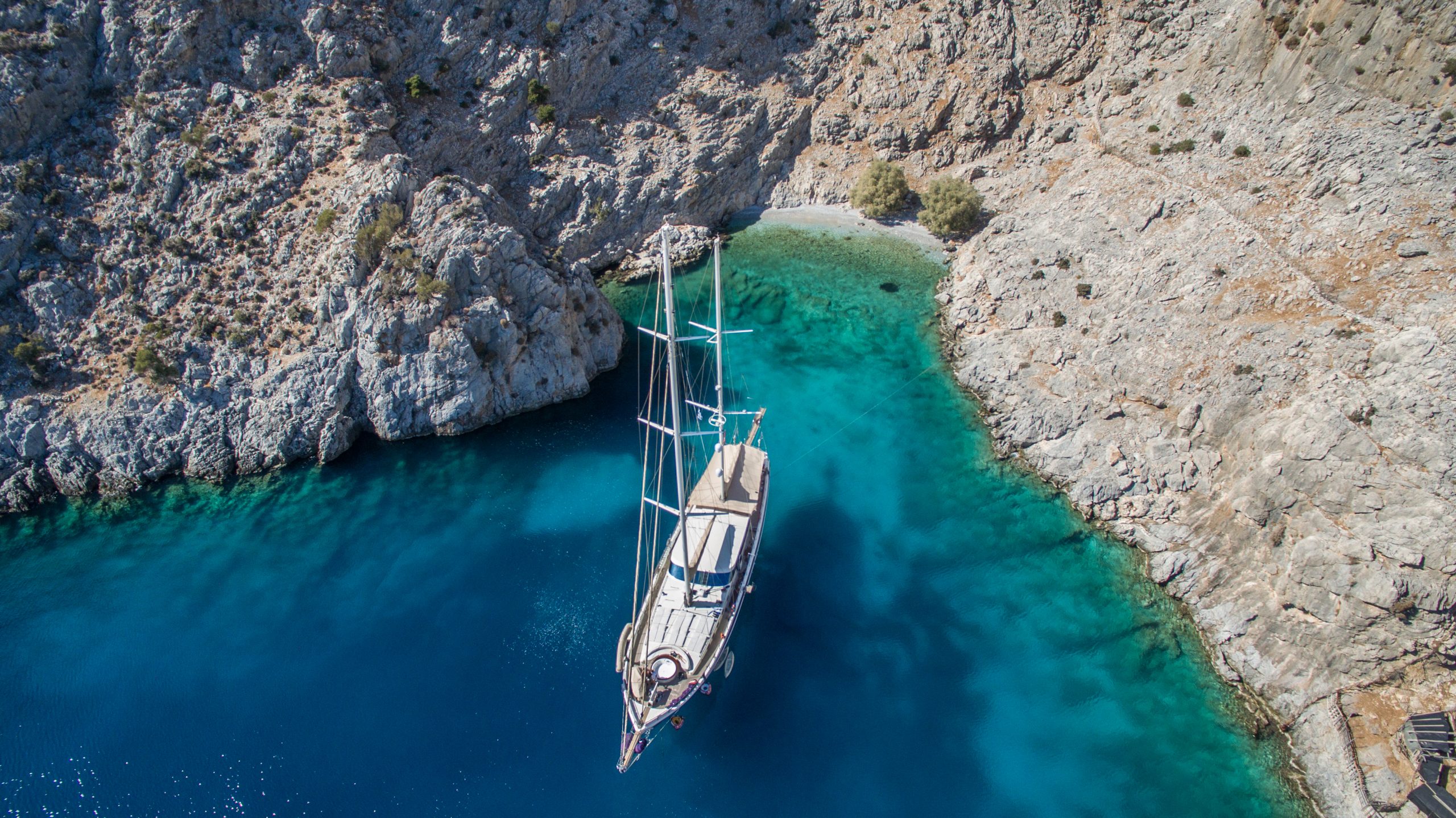 DOLCE MARE Aerial view of a sailboat anchored in clear turquoise water near a rocky coastline, with steep cliffs and a small sandy beach surrounded by rugged terrain.
