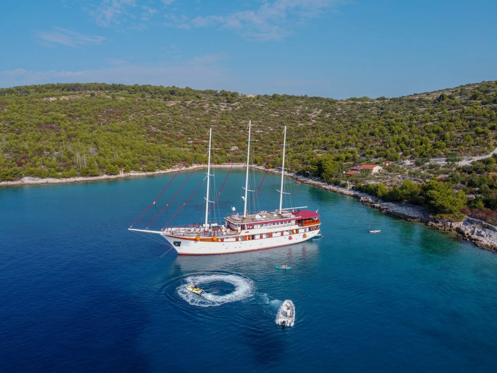 A large white sailing yacht is anchored in a calm, blue bay surrounded by green, tree-covered hills. A small motorboat and a person on a jet ski create a circular wake near the yacht. The sky is clear and sunny.