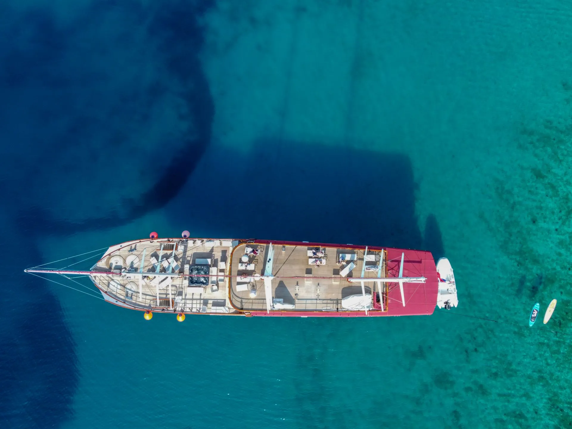 Aerial view of a large yacht with a red canopy anchored in clear blue water, with a small boat and two paddleboarders nearby.