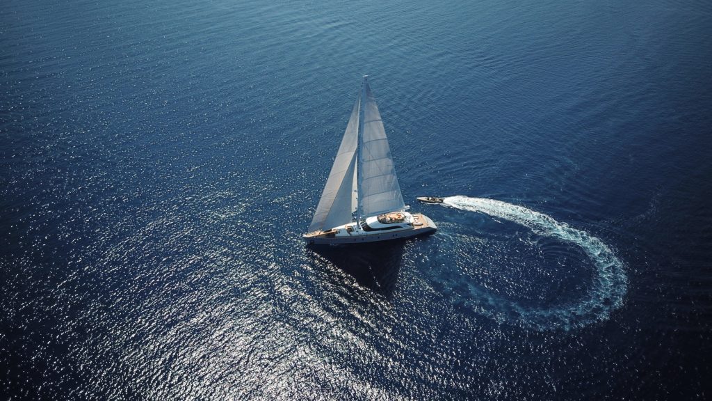 Aerial view of a sailboat gliding on calm blue water, with a small motorboat creating a circular trail nearby, both surrounded by shimmering sunlight reflections.