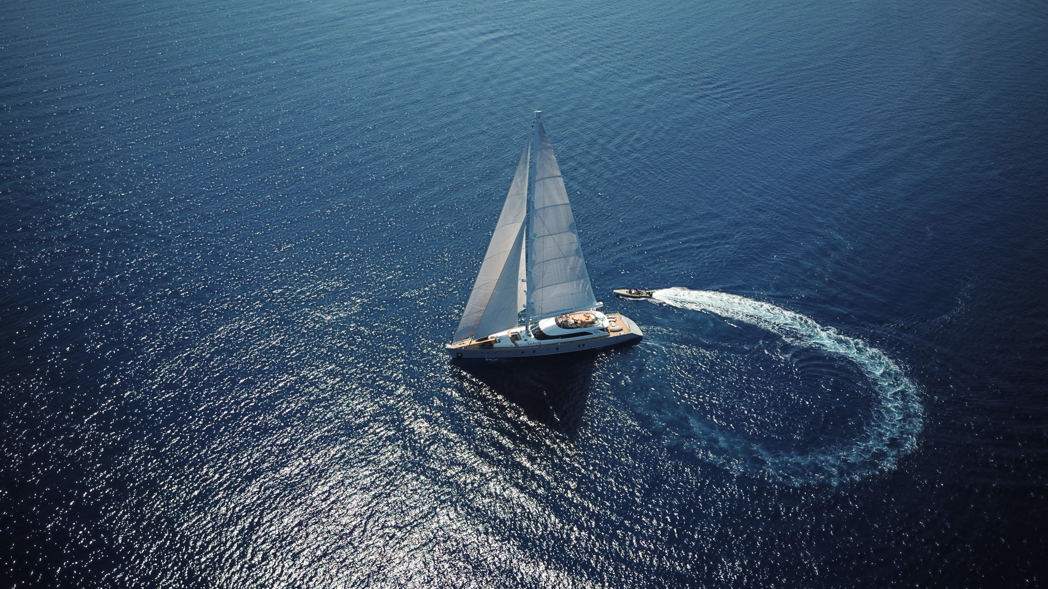 Aerial view of a sailboat gliding on calm blue water, with a small motorboat creating a circular trail nearby, both surrounded by shimmering sunlight reflections.