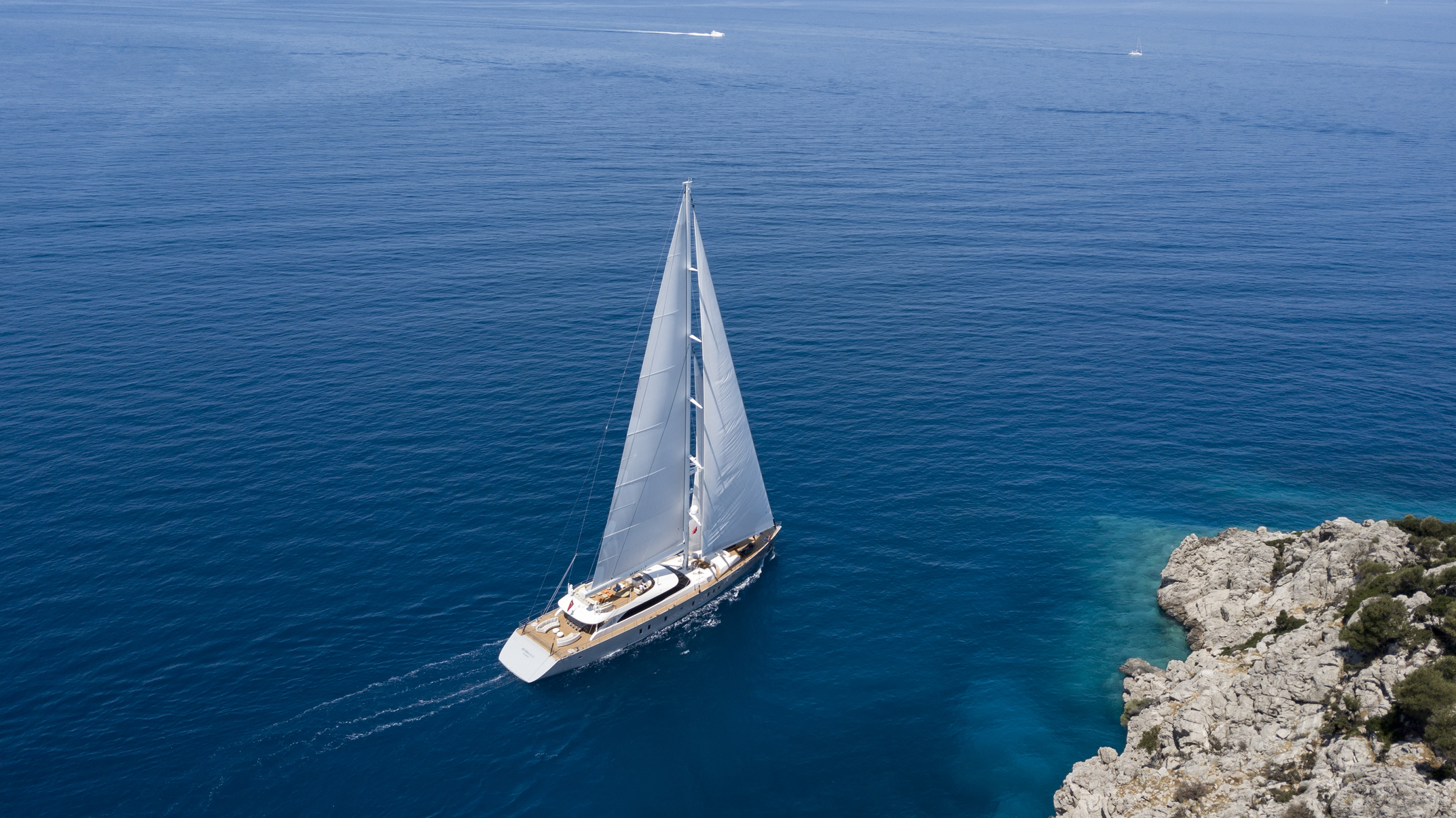 A white sailboat with raised sails glides across calm, deep blue water near a rocky coastline with clear turquoise shallows under a sunny sky.