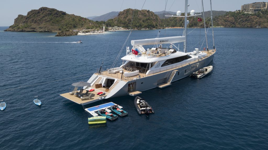 A luxury yacht anchored in clear blue water near a mountainous coastline, with jet skis, paddleboards, and a small boat tied alongside. The yacht has multiple decks and outdoor seating areas.