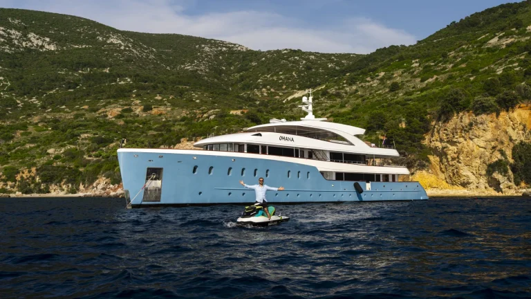 A person rides a jet ski on the water near a large, modern blue yacht anchored by a forested hillside under a partly cloudy sky.