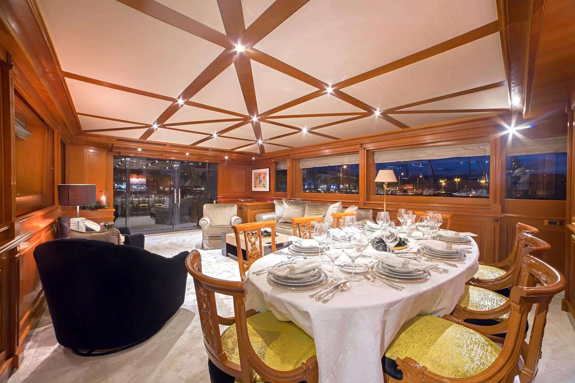 Elegant yacht dining room with a set table for eight, wooden chairs, plush seating area, large windows offering a marina view, and warm overhead lighting.