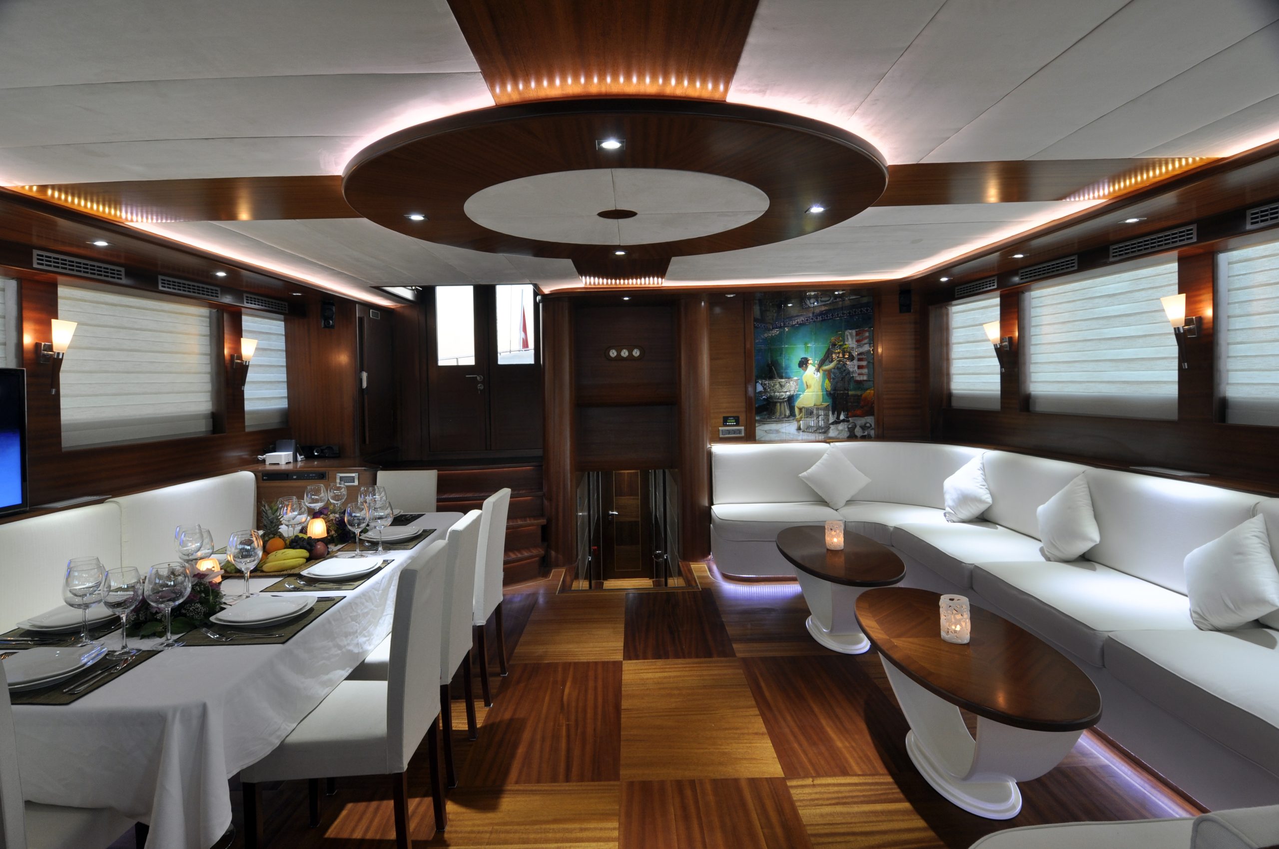 DOLCE MARE Luxurious yacht interior with warm wood paneling, white cushioned seating, a dining table set with glassware, candles on coffee tables, modern lighting, and large windows with blinds.