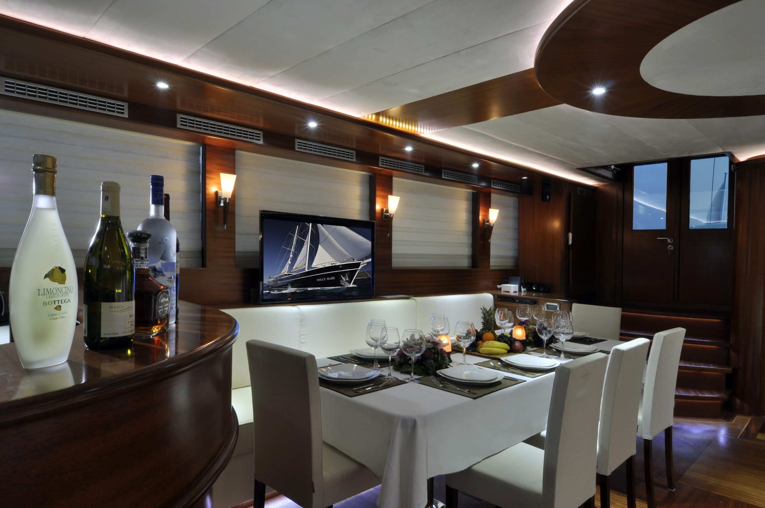 DOLCE MARE Elegant yacht dining area with a modern, wood-accented interior, set table for six, bottles and fruit on the counter, wall-mounted TV showing a sailboat, and steps leading to a doorway in the background.